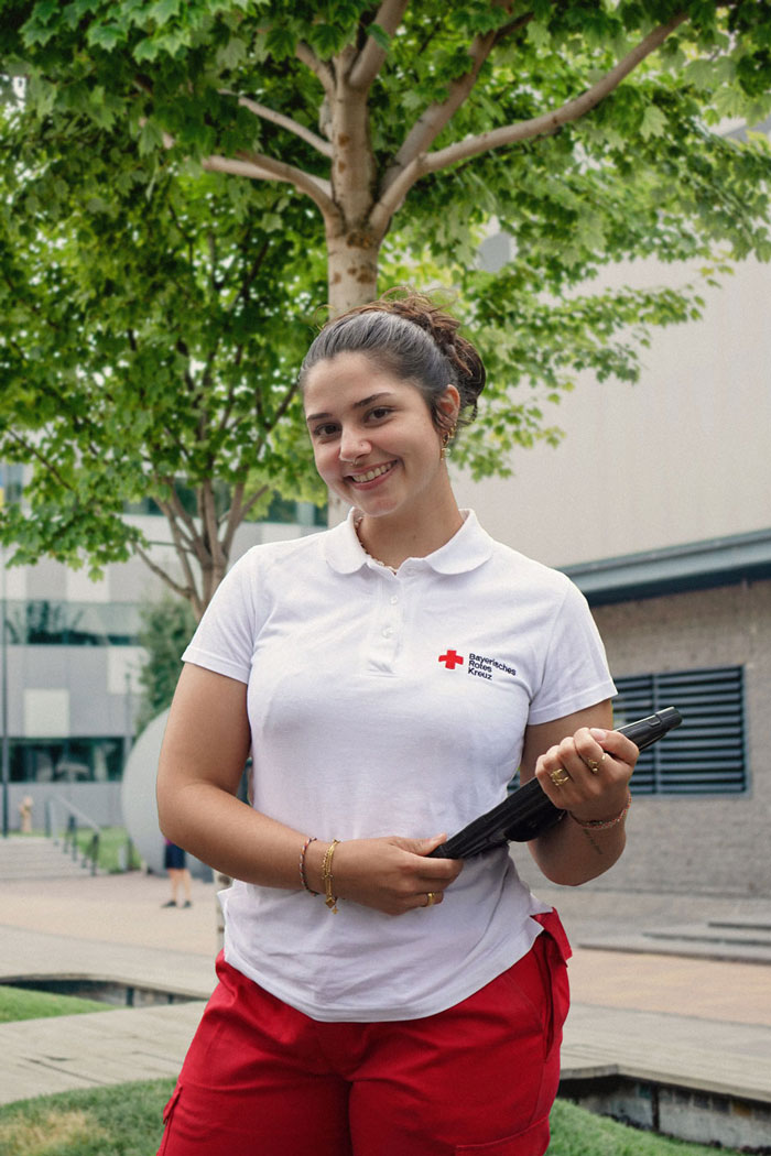 Eine junge Fundraiserin steht lächelnd in einem Wohngebiet und trägt die Kleidung des Bayerischen Roten Kreuzes. Sie hält ein Tablet in der Hand.