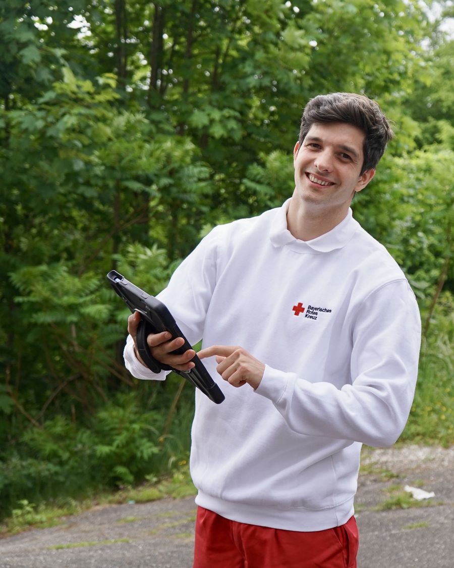 Ein Fundraiser im weißen Pullover des Bayerischen Roten Kreuzes zeigt lächelnd auf ein Tablet in seiner Hand.